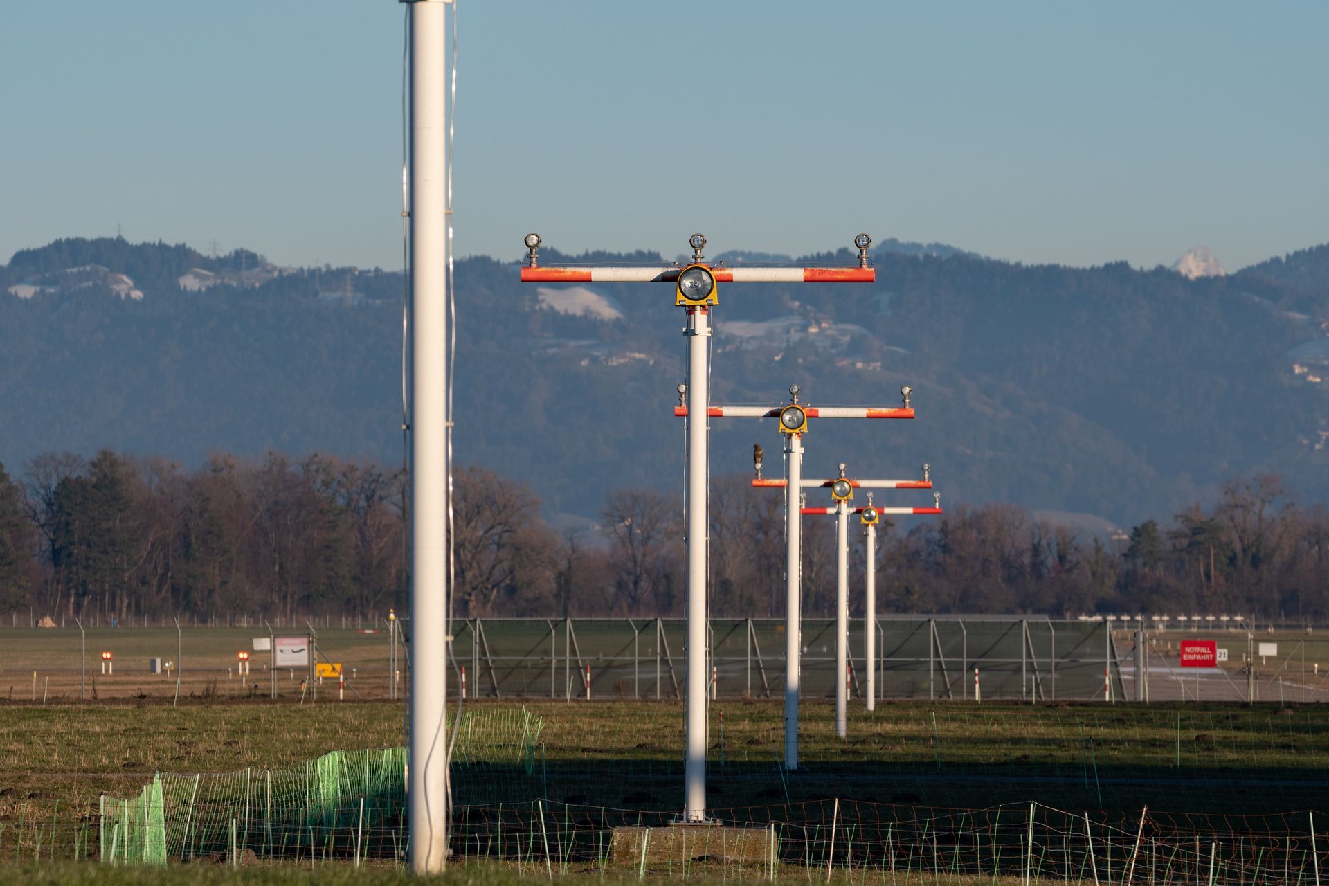 Airport approach lighting system, white poles with horizontal orange and white lights, green field, mountain background.