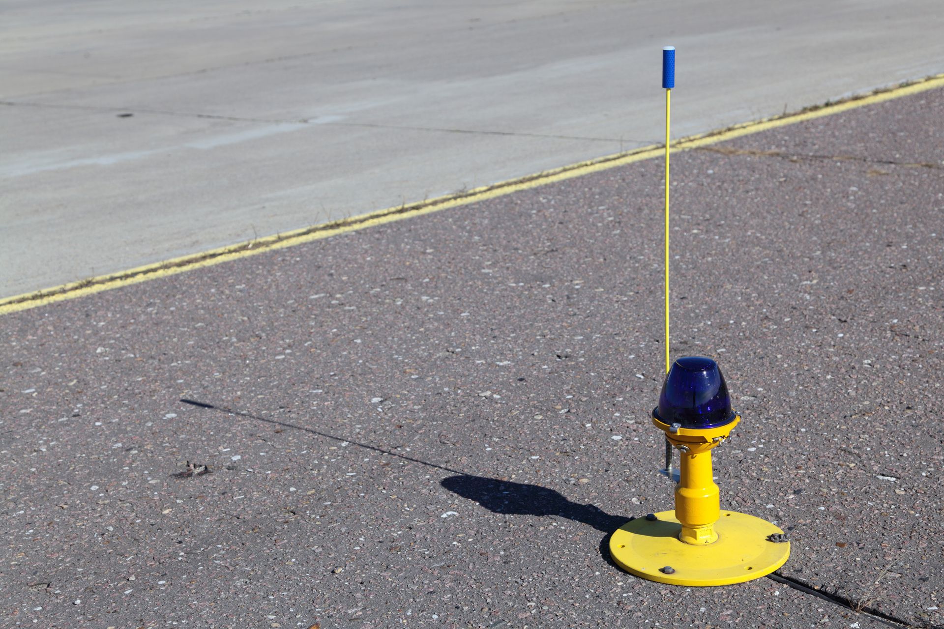 Yellow airport taxiway light with a blue lamp, casting a shadow on the asphalt.