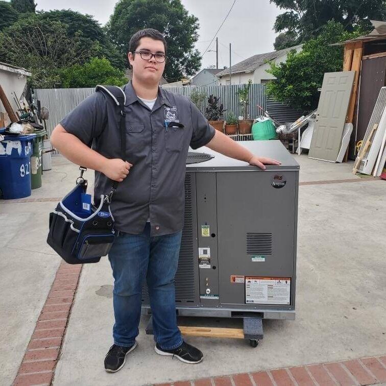 Man in grey work shirt poses next to a generator, holding a tool bag. Outdoors, sunny.