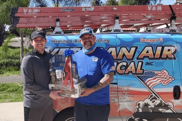 Two men holding a gift basket in front of a Veteran Air Mechanical Inc. van.