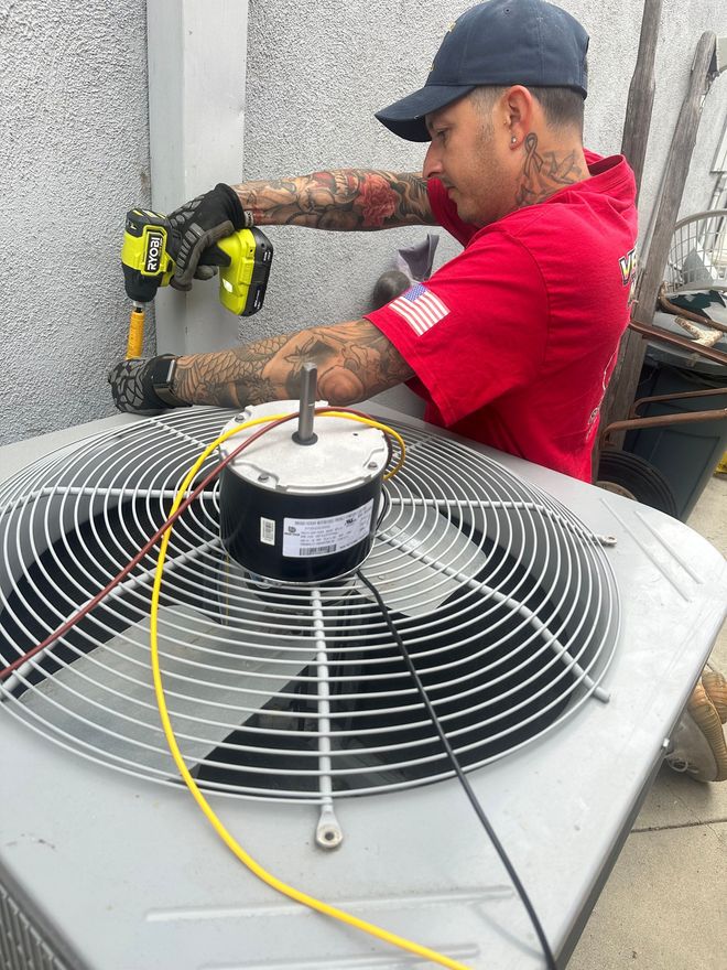 Man in red shirt works on an air conditioning unit outside, using a power drill.