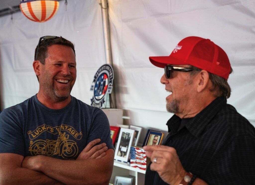 Two men laughing; one in a red cap and sunglasses gestures while the other has arms crossed. Inside a tent.