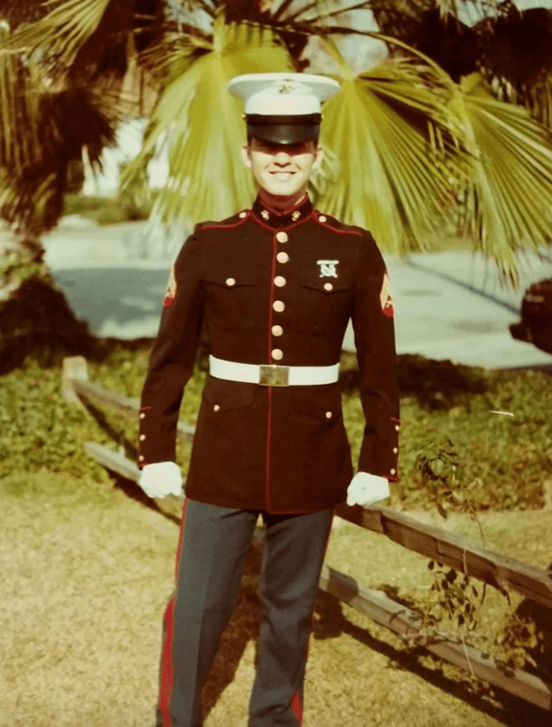 Marine in dress blues standing outdoors near a wooden fence and palm trees.