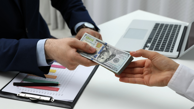 Person in suit handing cash to another person at a desk with a laptop and financial documents.
