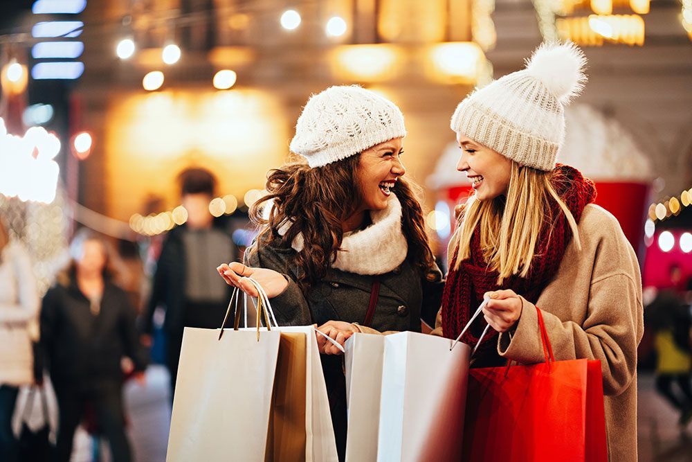Two women, wearing winter hats and coats, laugh while holding shopping bags on a festive street at night.
