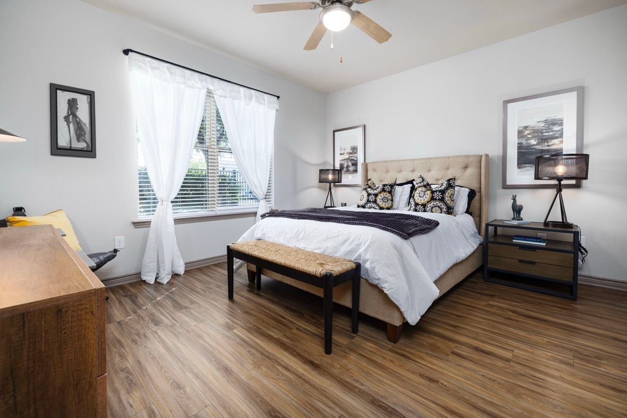 Bedroom with a tufted beige headboard, two nightstands, bench at foot of bed, and a window with white curtains.