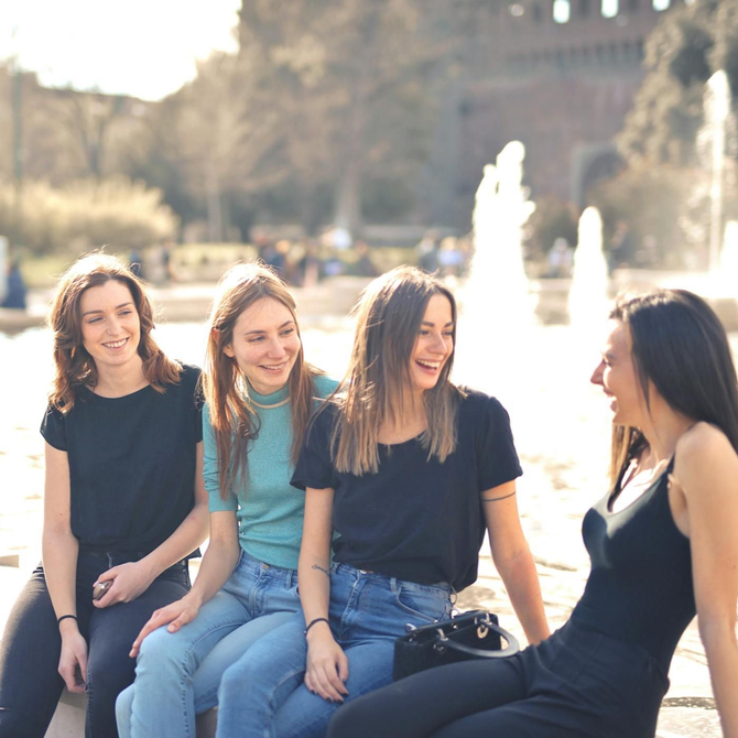 Four friends sit together and laugh near a sunny outdoor fountain, dressed in casual tops and jeans.