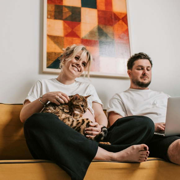 A smiling person pets a cat while sitting on a yellow sofa next to another person looking at a laptop.