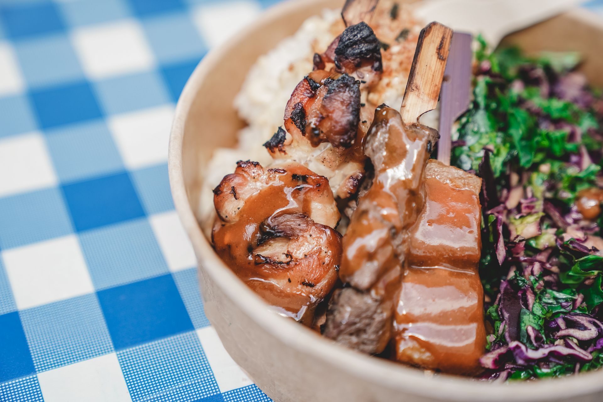 Bol garni de viandes grillées, de riz et de salade, posé sur une nappe à carreaux bleus et blancs.