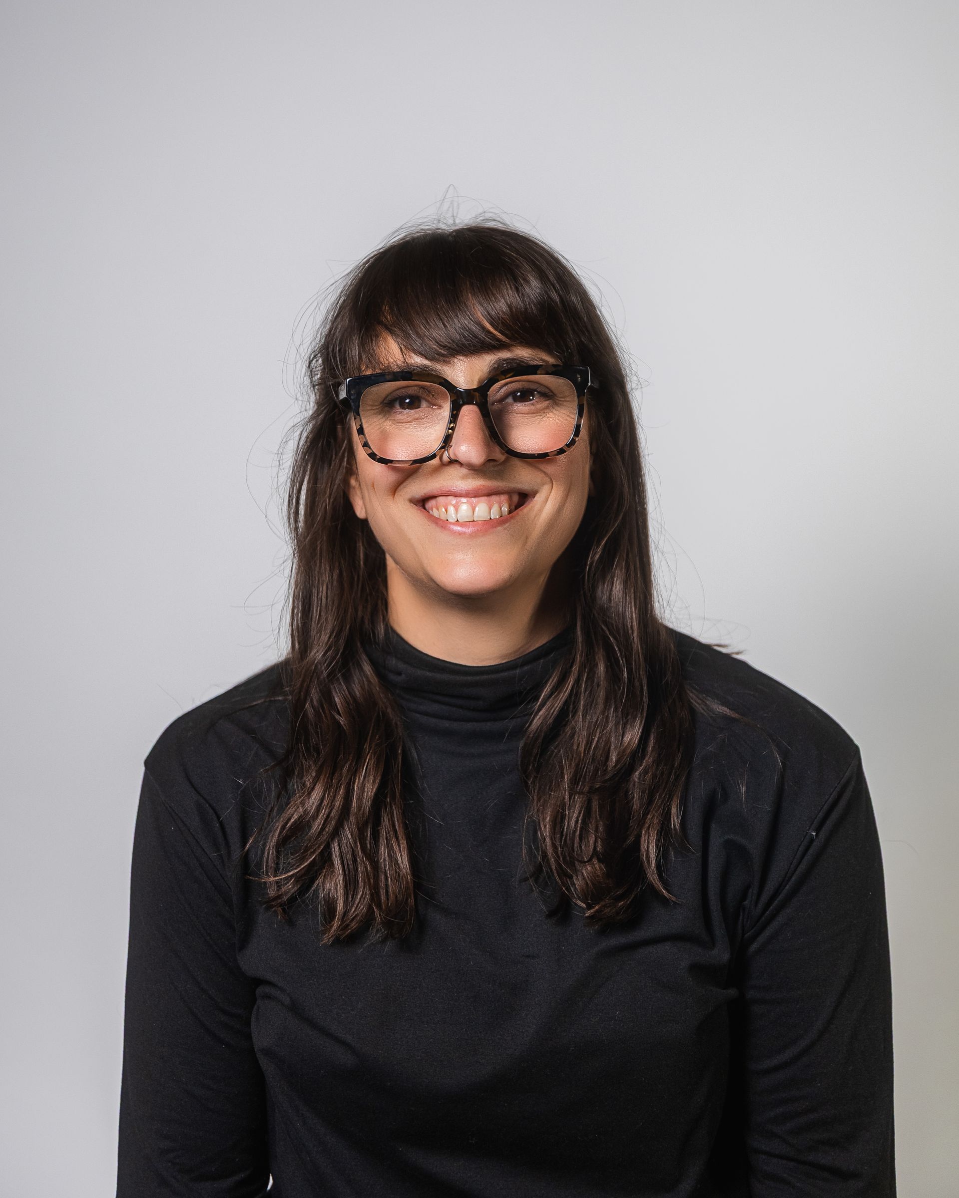 Une femme aux cheveux foncés et portant des lunettes sourit, vêtue d'un col roulé noir.