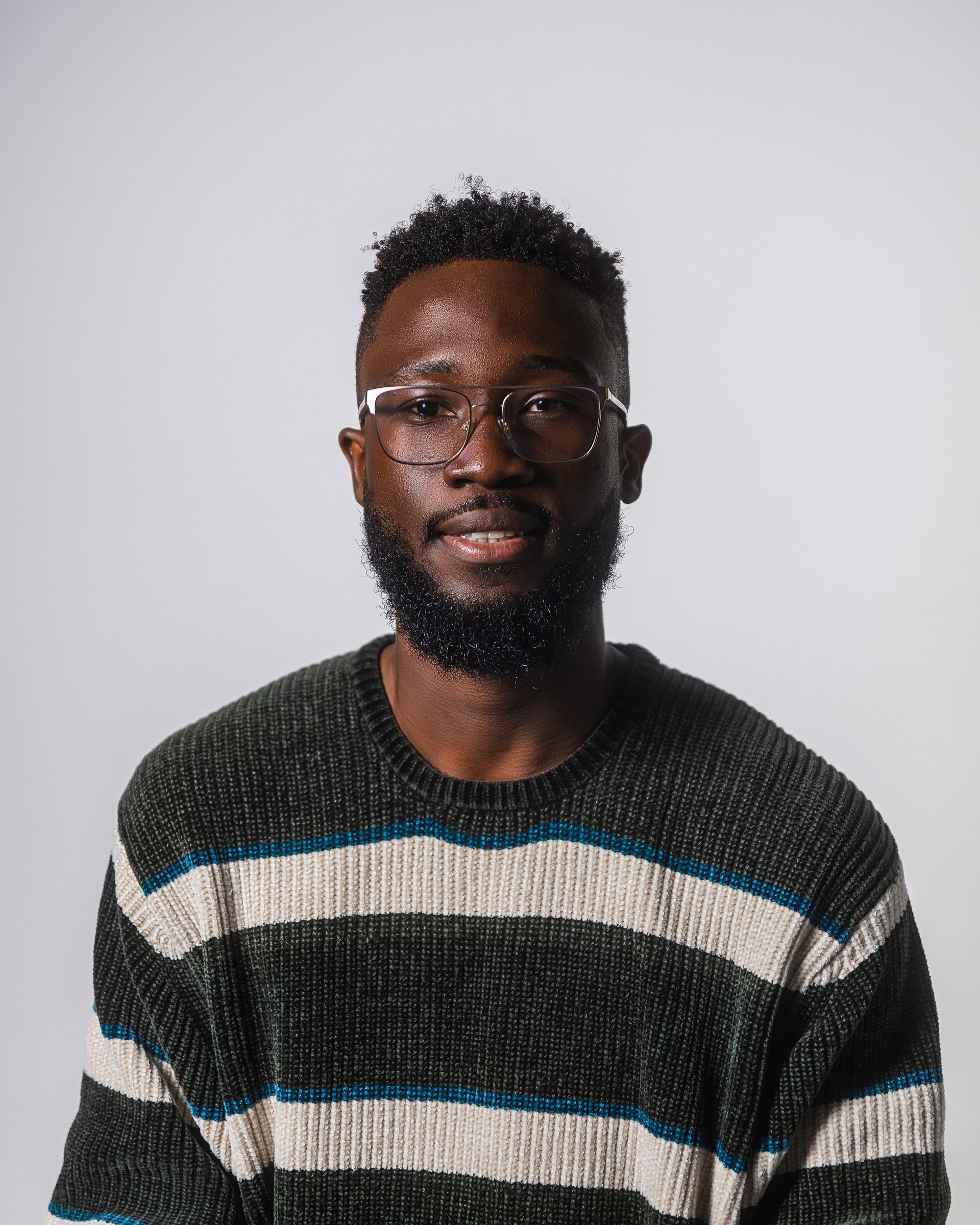 Un homme portant des lunettes et une barbe, vêtu d'un pull rayé, sourit sur un fond blanc.