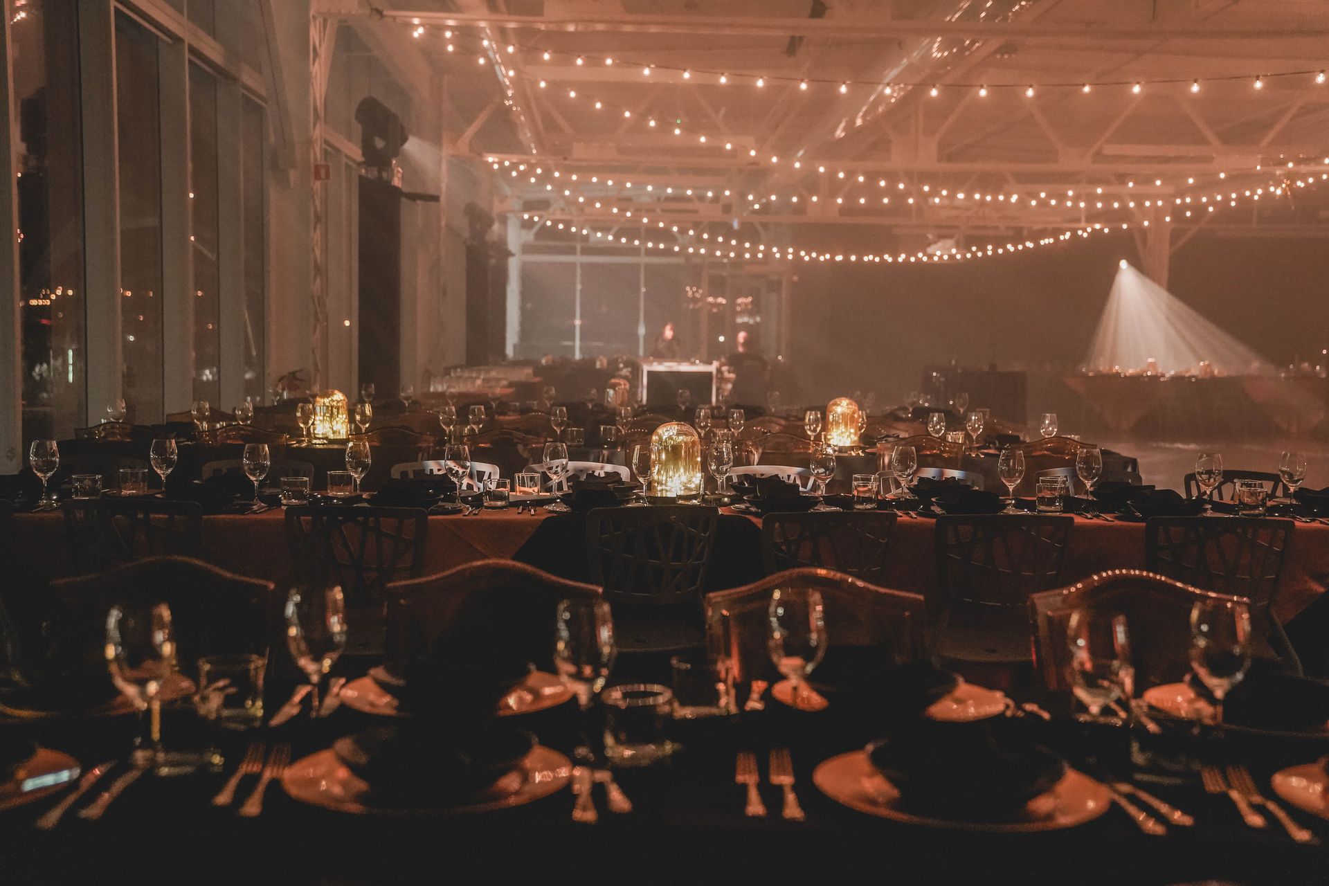Des tables à manger formelles sont dressées dans une pièce brumeuse, éclairée par des guirlandes lumineuses et des spots à lumière chaude.