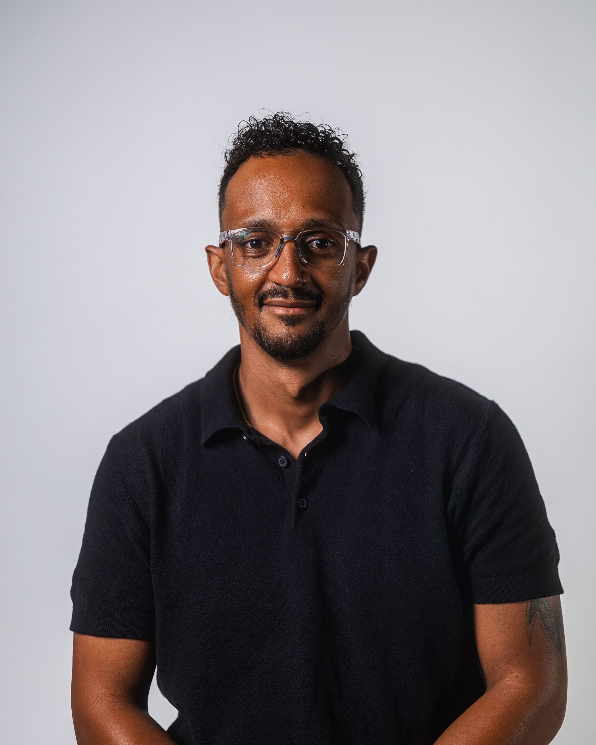 Un homme portant des lunettes et un polo noir sourit. Fond clair.