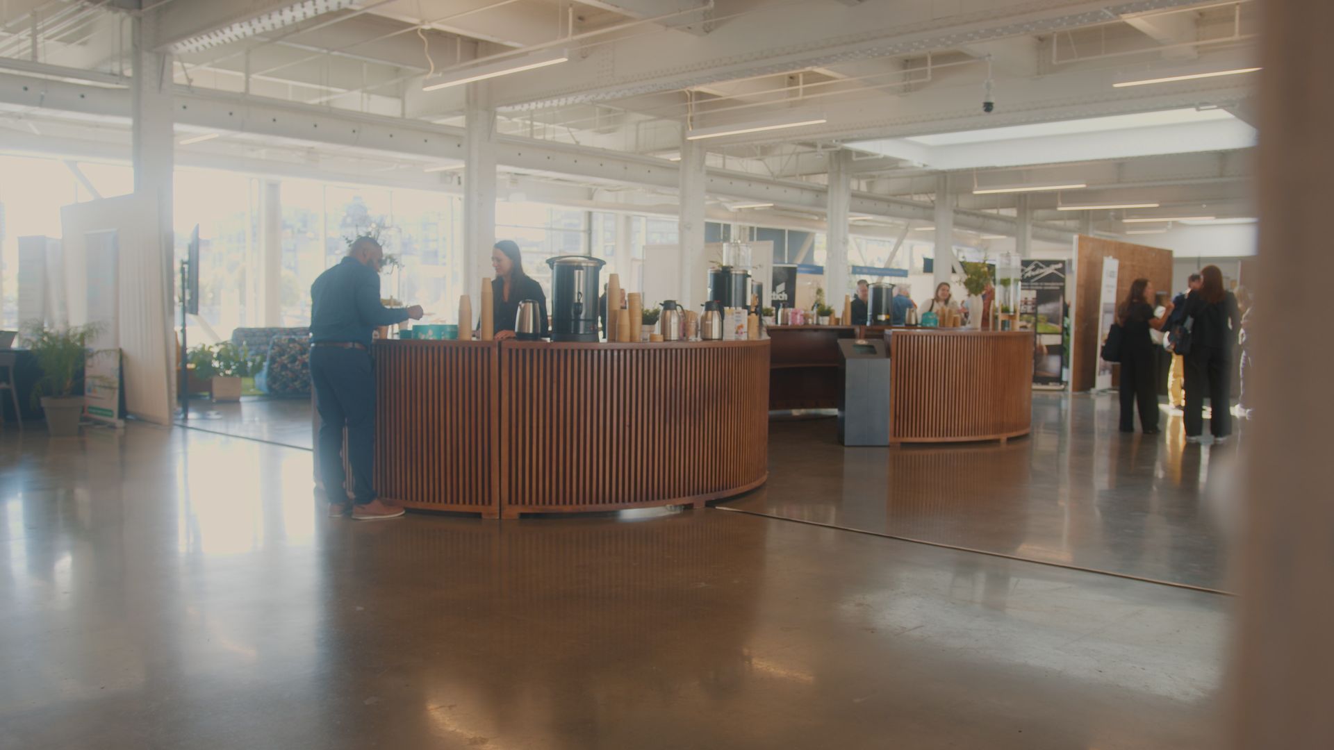 Pièce spacieuse avec des personnes près de tables en bois. Certaines se tiennent derrière les tables, d'autres circulent à proximité. Sol gris et plafond blanc.