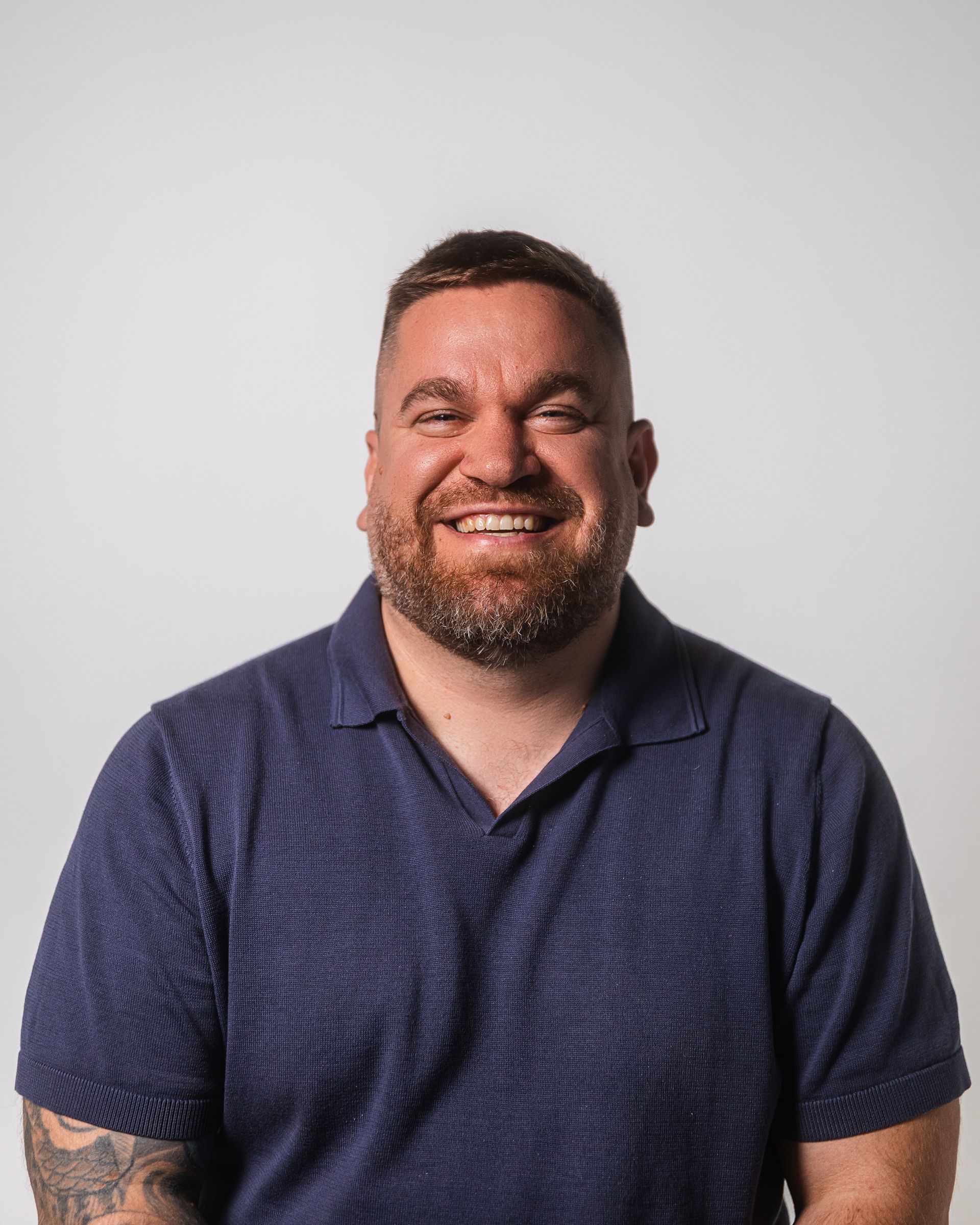 Homme barbu souriant, portant un polo bleu, sur fond blanc.
