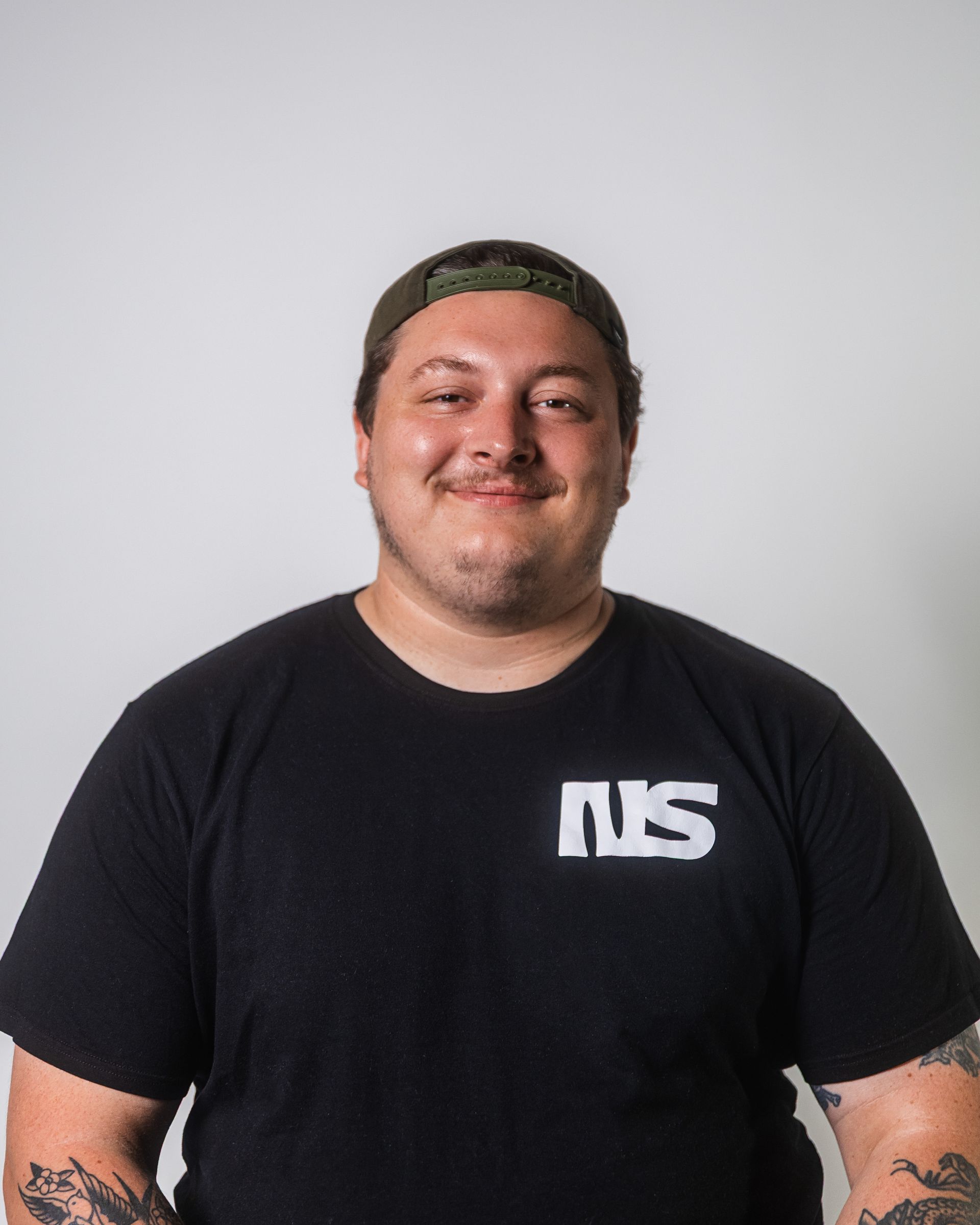 Un homme portant un t-shirt noir et une casquette verte sourit devant un fond blanc.