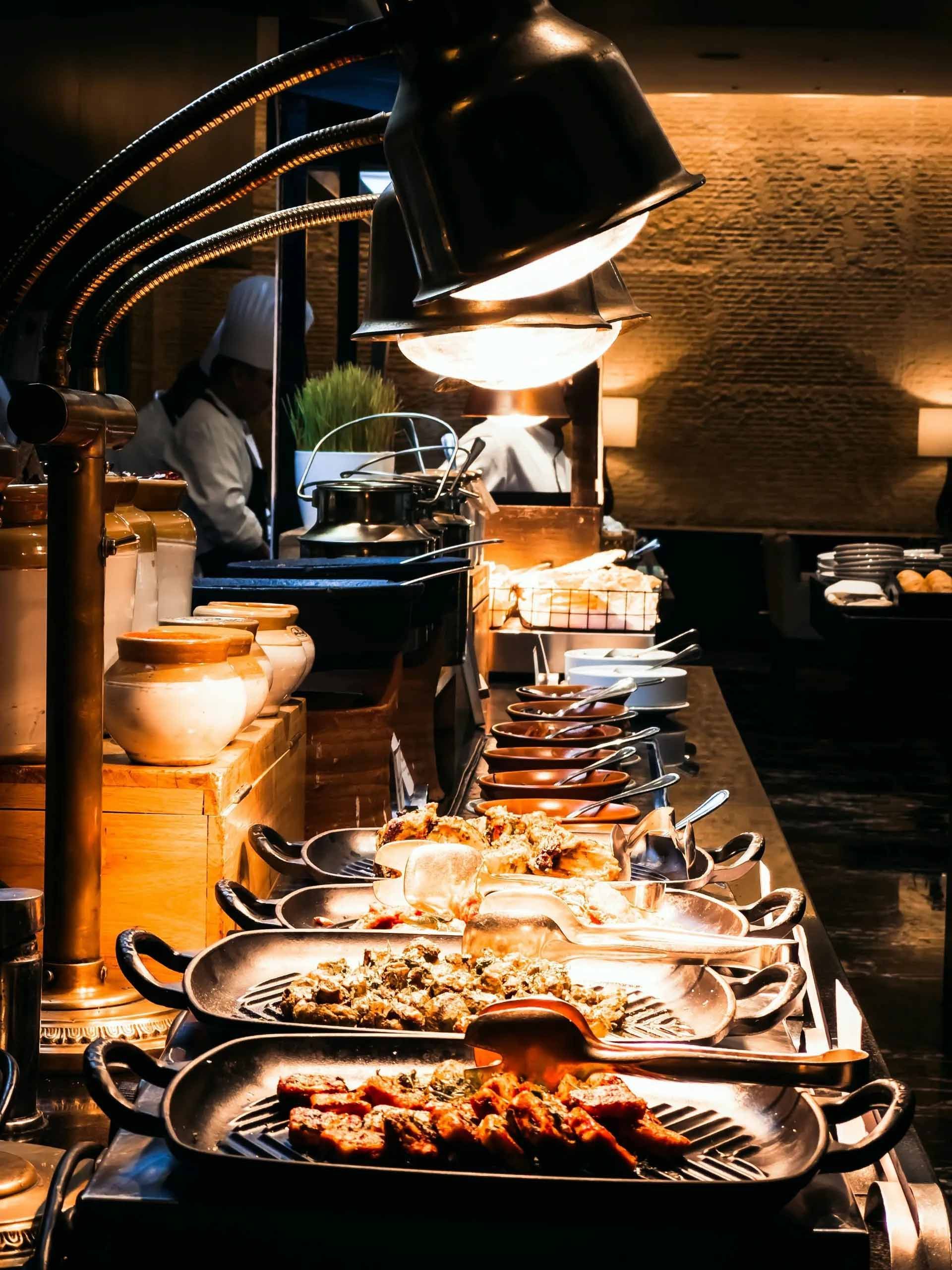 Buffet composé de plusieurs plateaux de nourriture, sous une lumière chaude, dans un restaurant à l'éclairage tamisé.
