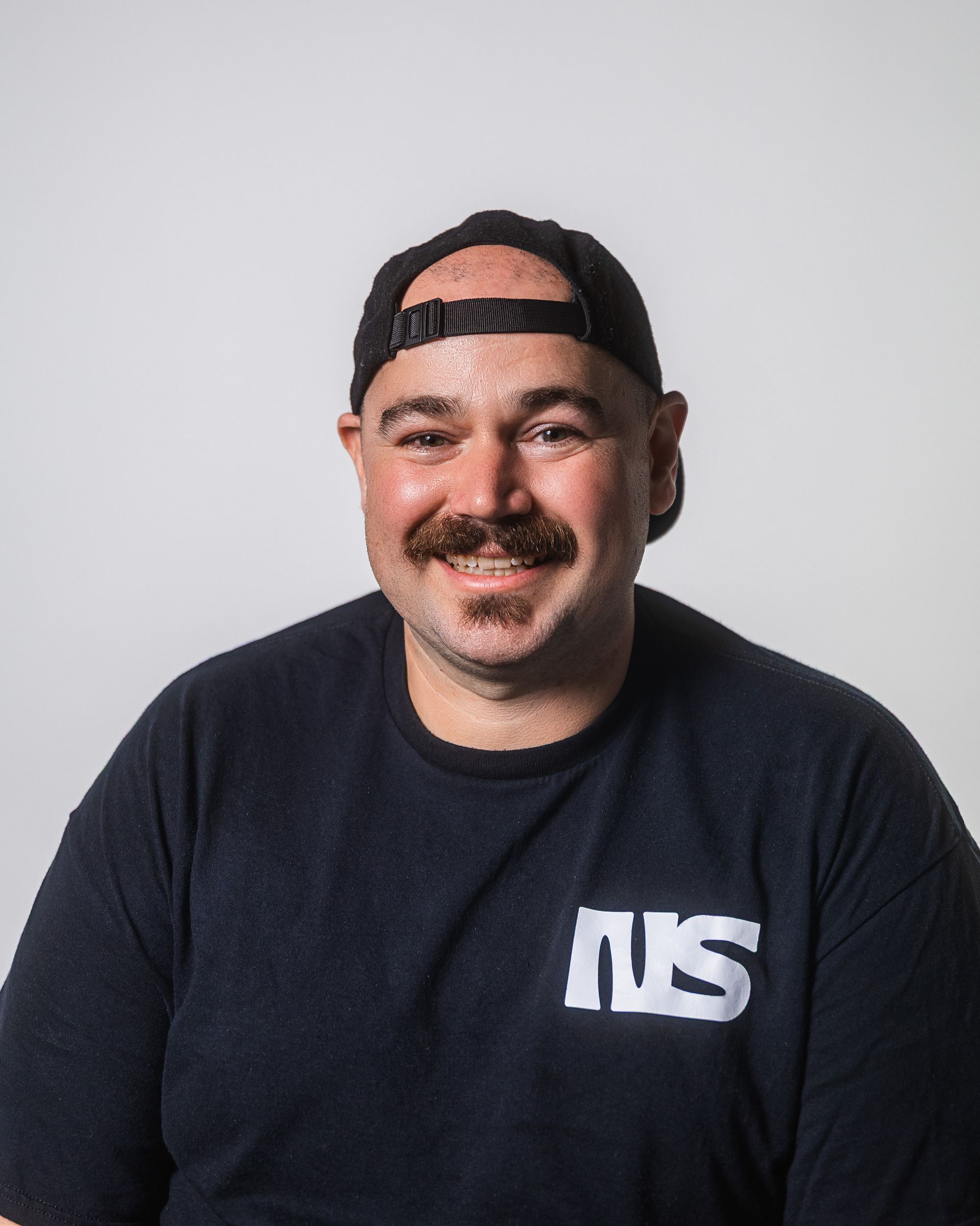 Un homme à moustache, portant une casquette noire et un t-shirt avec l'inscription « NWS », sourit.
