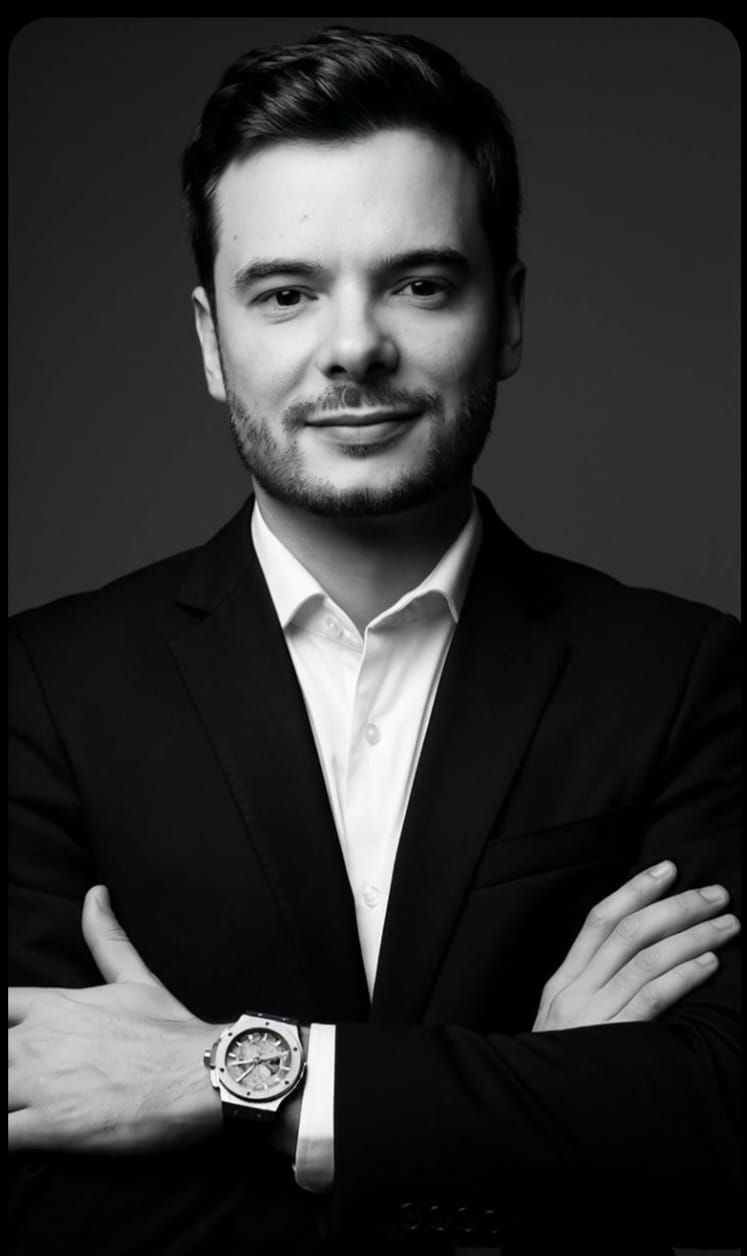 Una persona con cabello corto y castaño y vello facial claro viste una camisa abotonada de color azul oscuro con puntos blancos frente a una pared de ladrillos.