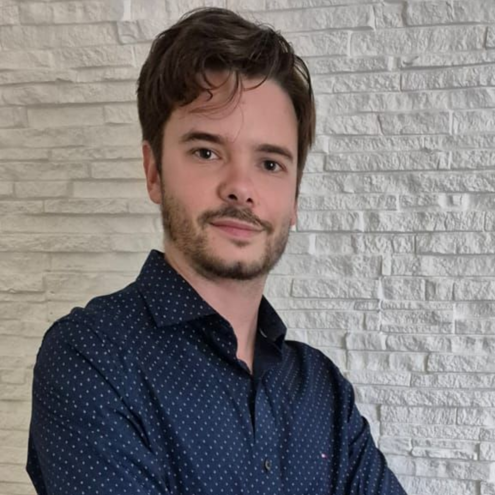 Una persona con cabello corto y castaño y vello facial claro viste una camisa abotonada de color azul oscuro con puntos blancos frente a una pared de ladrillos.