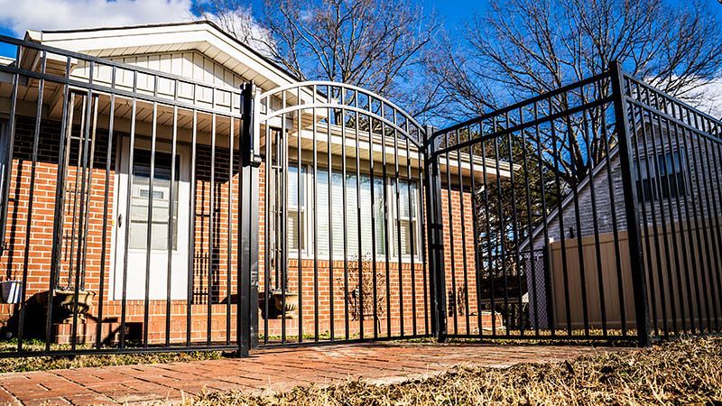 A black metal fence and arched gate stand in front of a brick house with a white door on a sunny day.