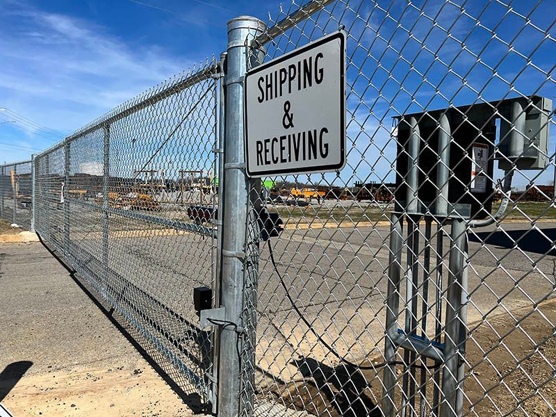 A chain-link gate and fence with a metal sign reading "SHIPPING & RECEIVING" under a bright blue sky.