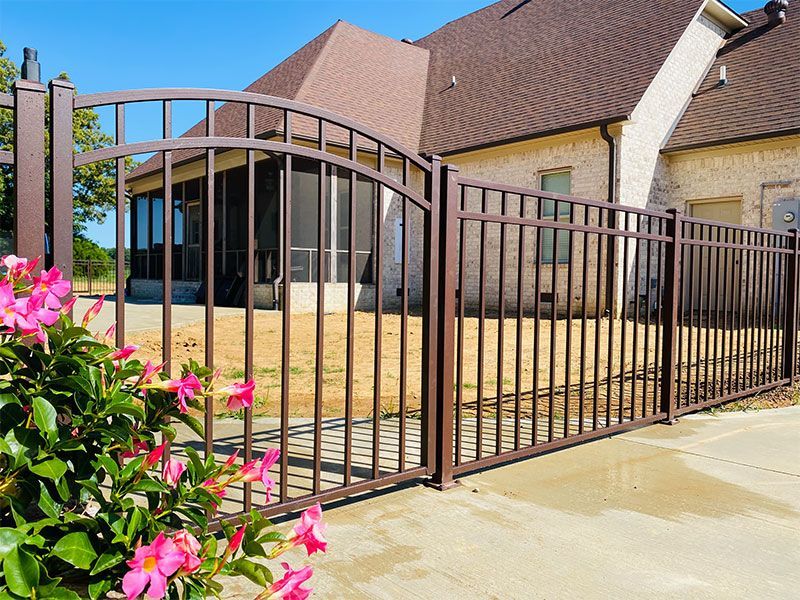 Brown metal gate and fence in front of a brick house with pink flowers at the left edge