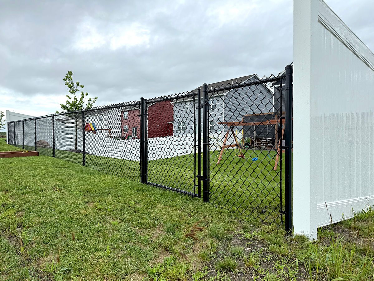 A black chain-link fence separates a grassy backyard with a play set from a neighboring yard with a white privacy fence.