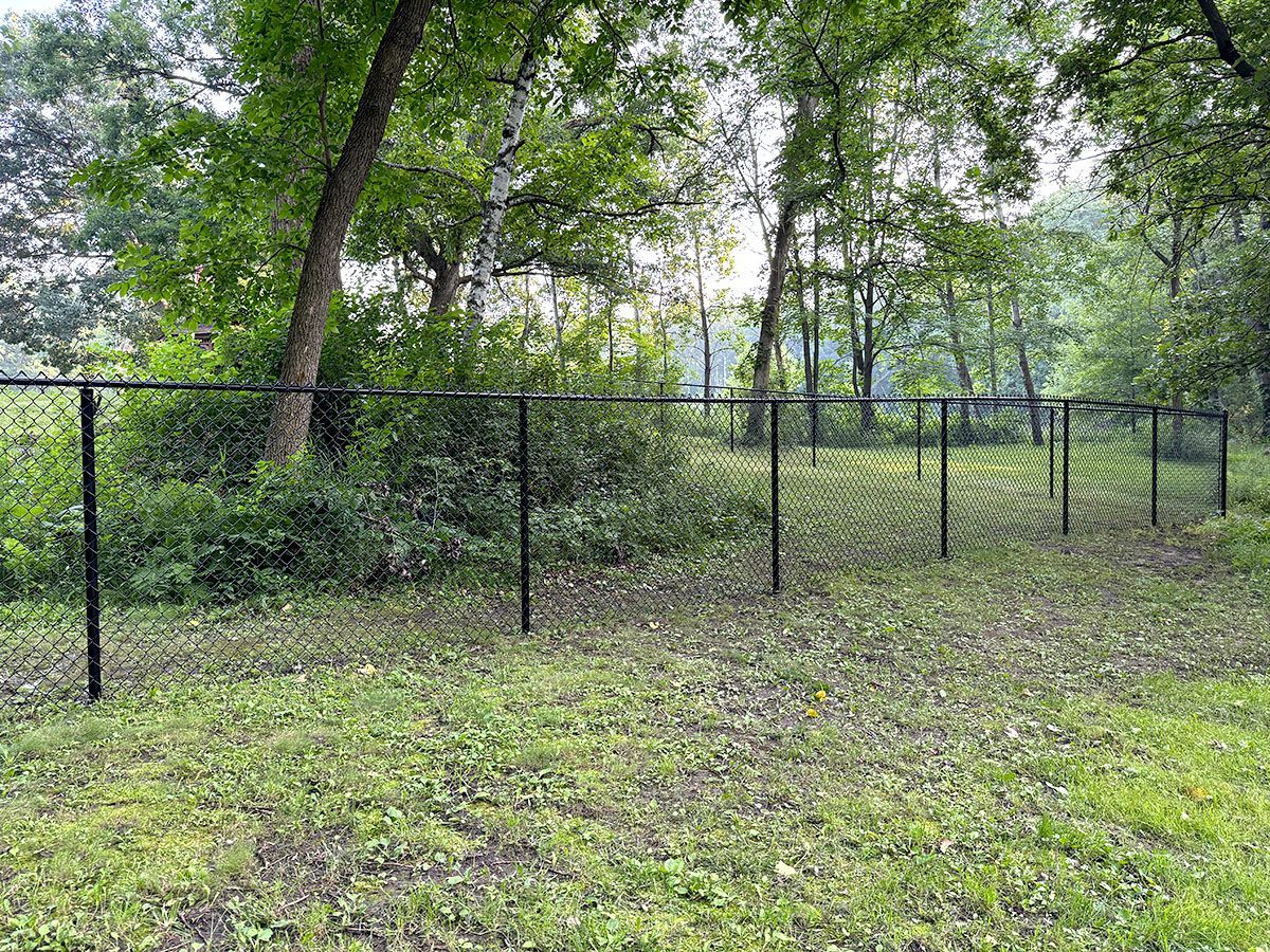 A black chain-link fence curves through a grassy area leading toward a wooded forest.