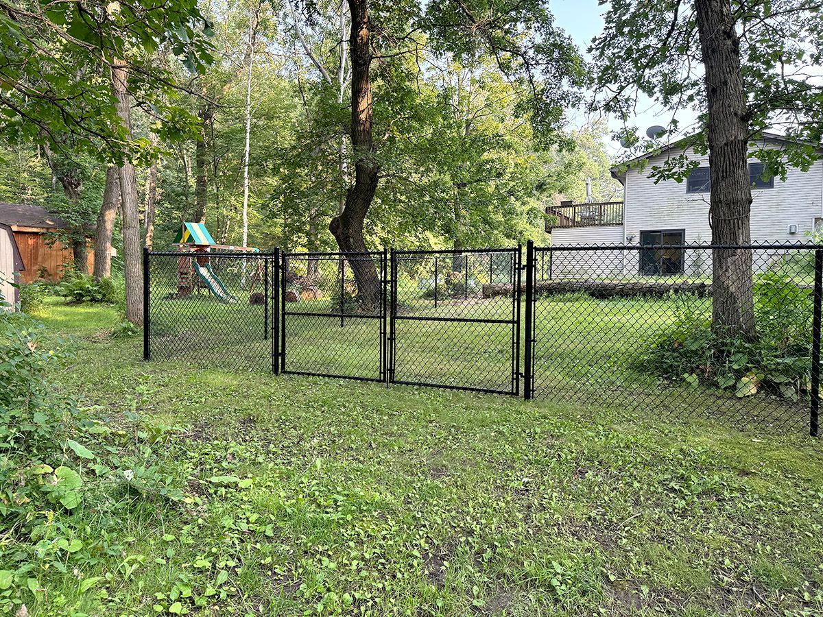 A black chain-link fence with a central gate in a grassy backyard, with trees and a house in the background.