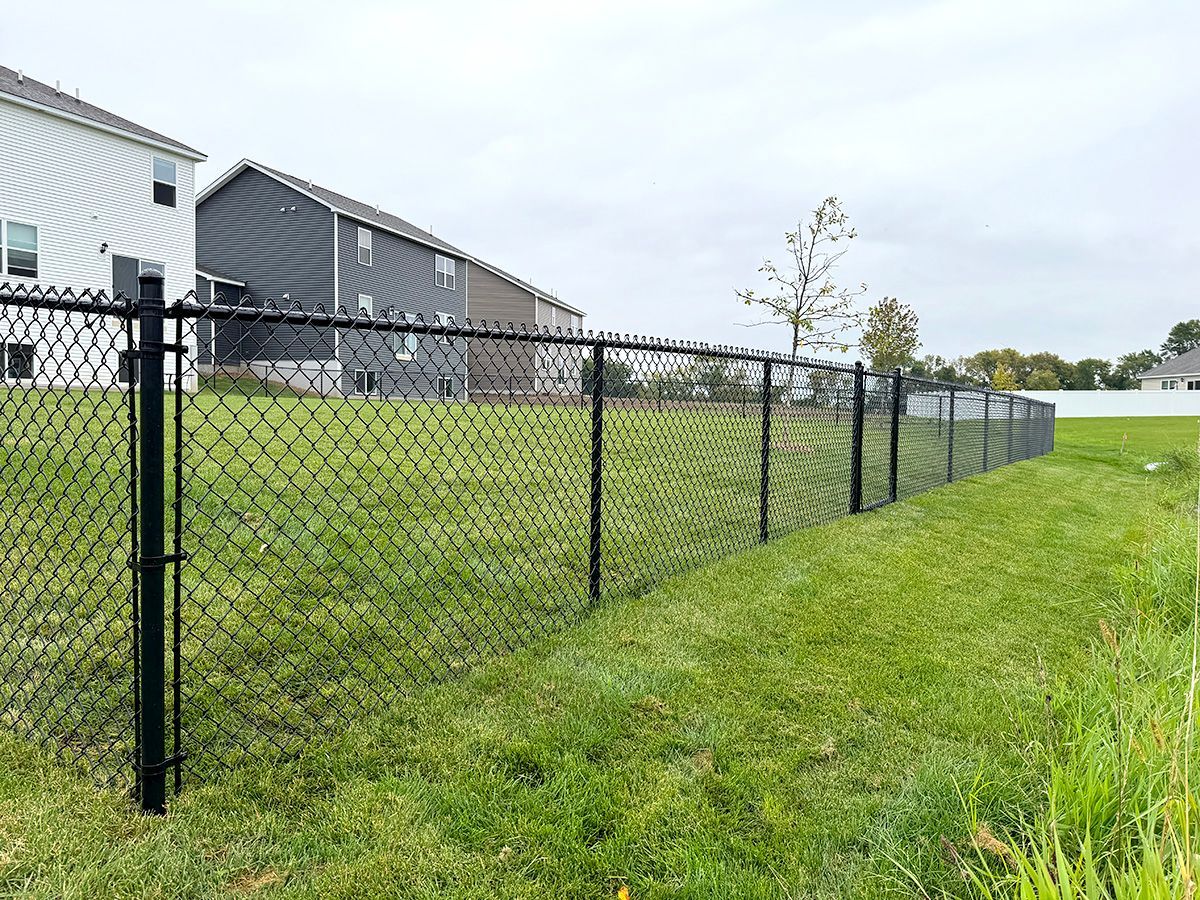 A black chain-link fence lines a green grassy yard in front of two suburban houses under a cloudy sky.