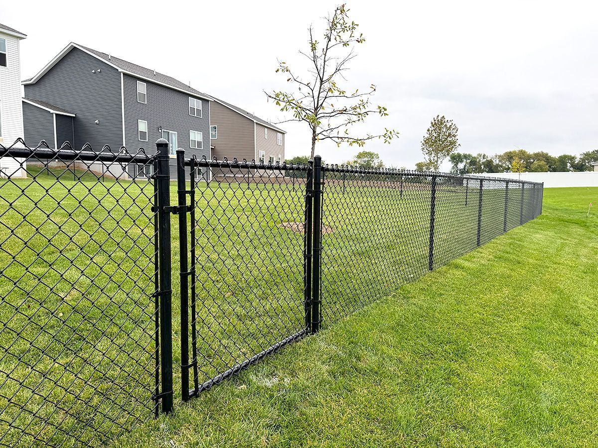 A black chain-link fence with an integrated gate stands on a grassy lawn in front of houses on a cloudy day.