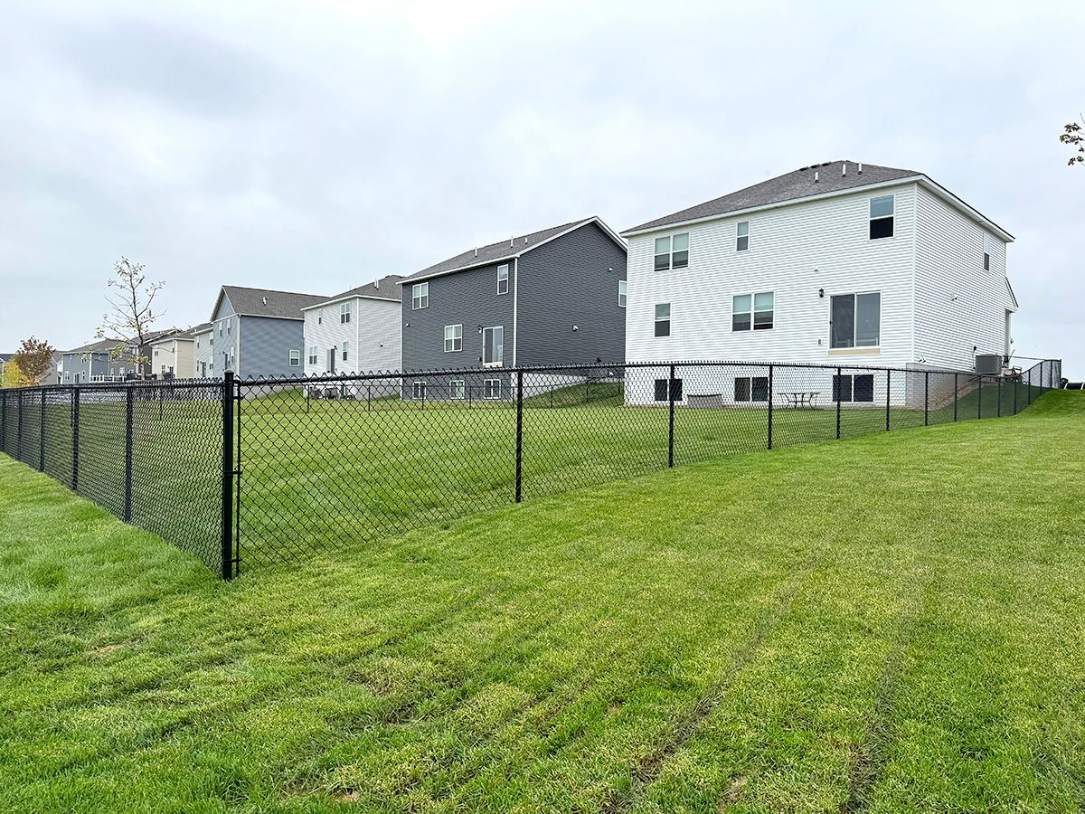 A black chain-link fence encloses a green grassy yard in front of a row of modern suburban houses under a cloudy sky.