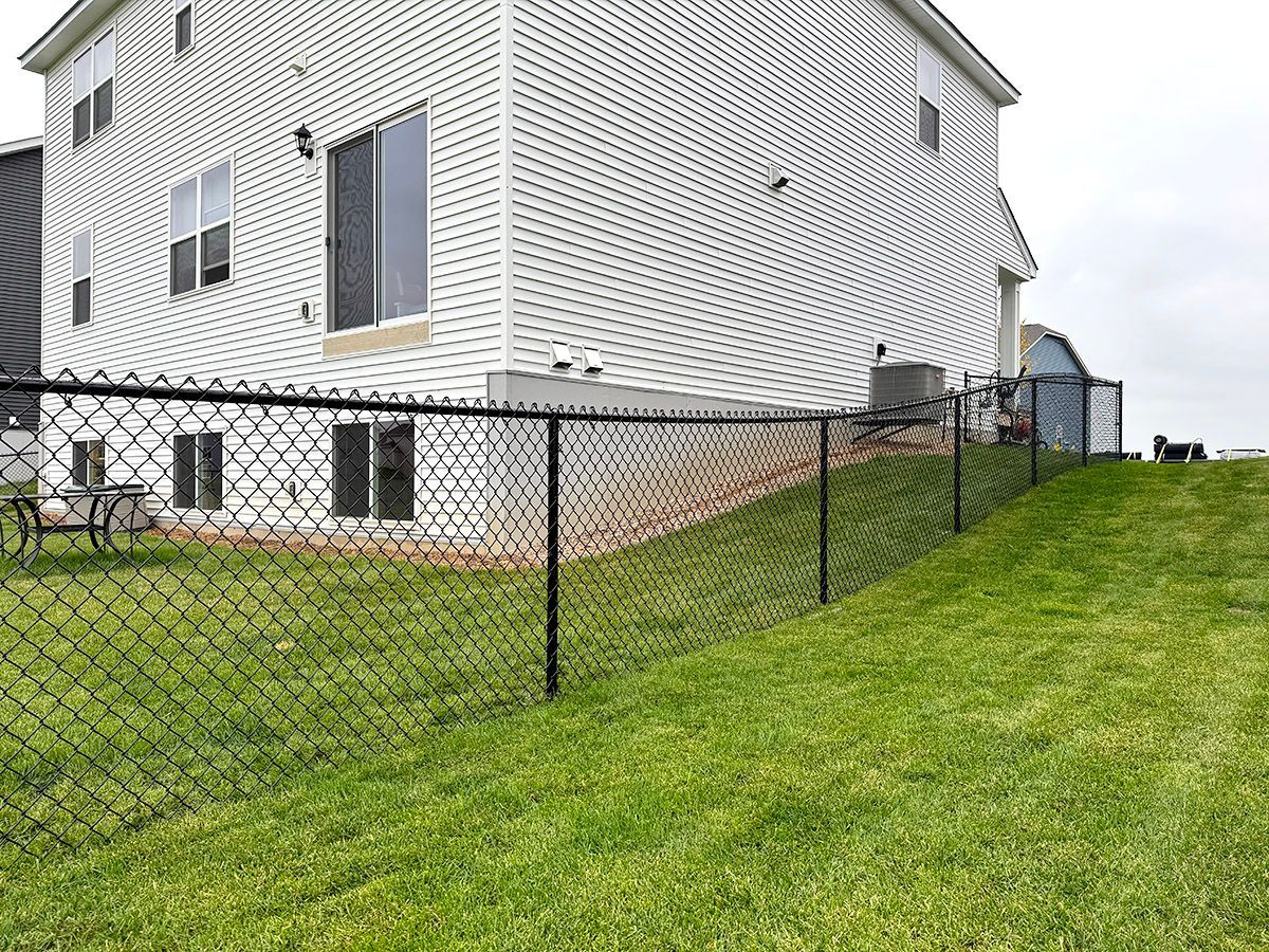 A white house with a sloped lawn is bordered by a black chain-link fence on a cloudy day.