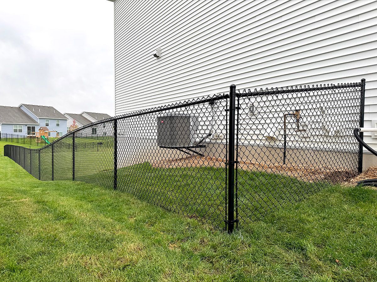 Black chain-link fence bordering a backyard lawn next to the side of a light-colored house with an AC unit.