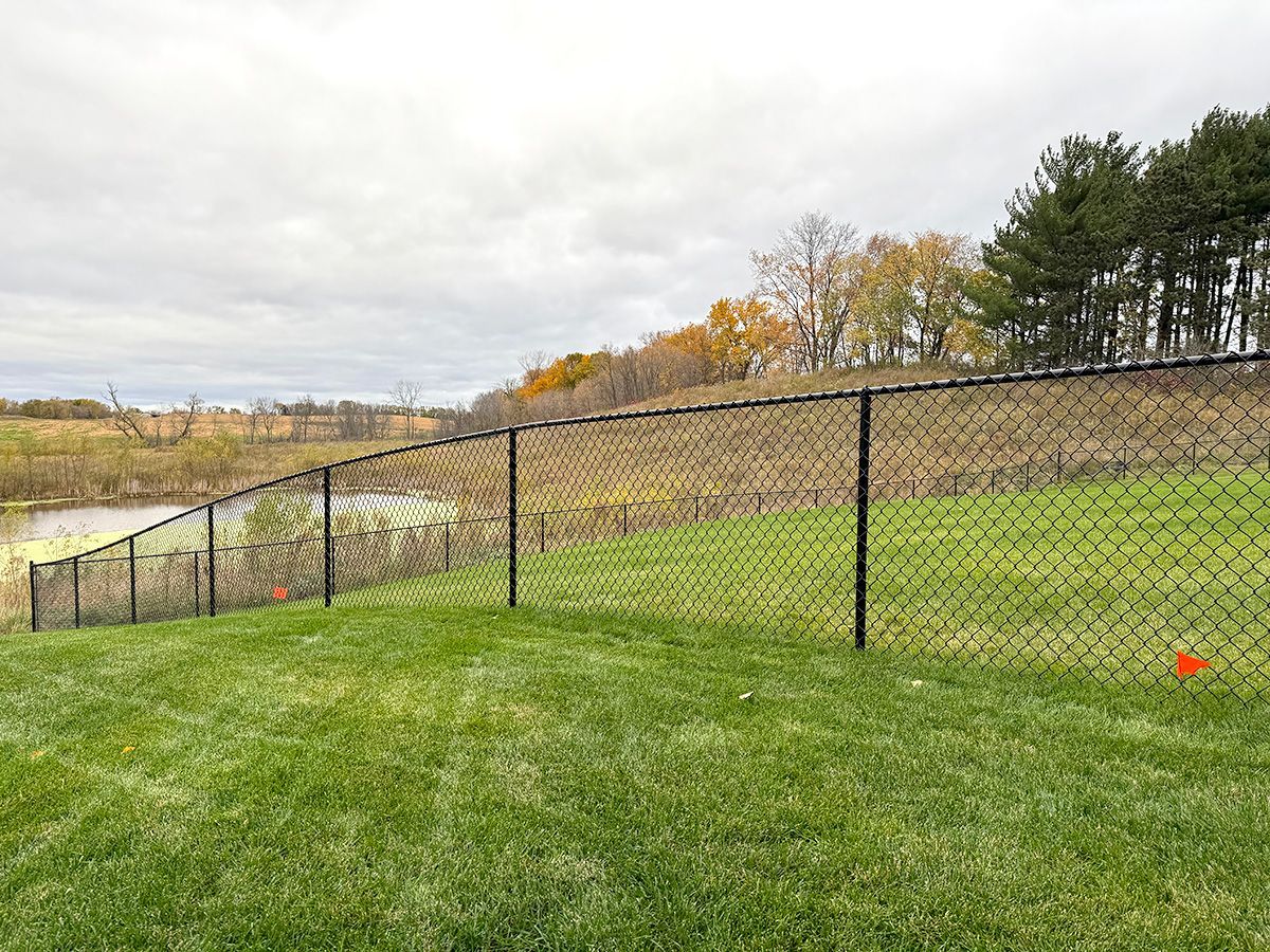 A black chain-link fence lines a sloping green lawn, with a body of water and autumn trees in the background.
