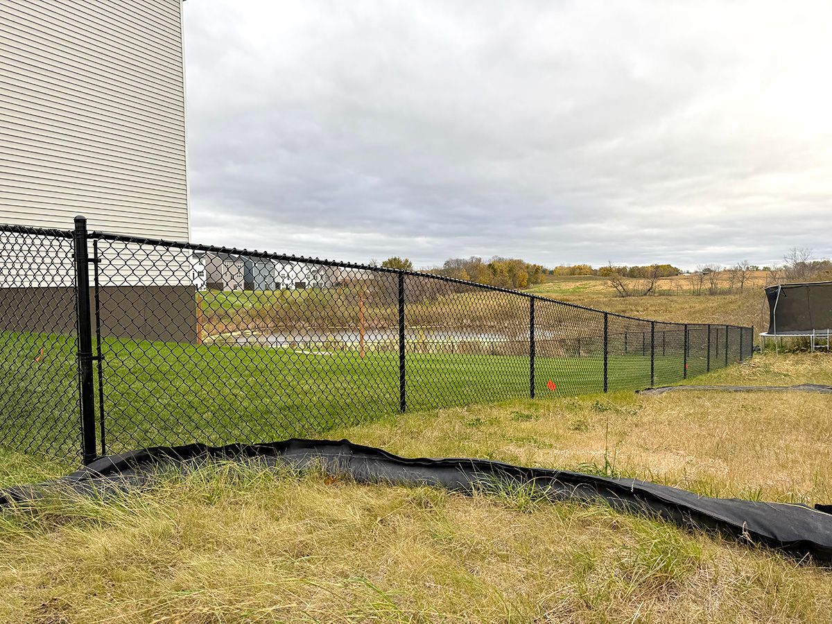 A black chain-link fence runs along a sloping grassy yard next to a beige building under a cloudy sky.