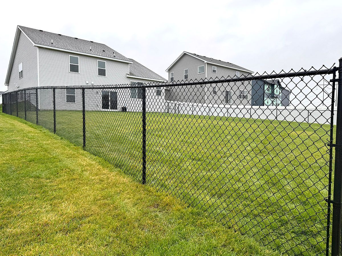 A black chain-link fence encloses a green grassy lawn in front of two gray houses under a cloudy sky.
