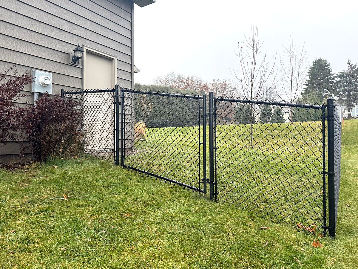 A black chain-link fence and gate installed on a grassy slope next to the side of a grey house.