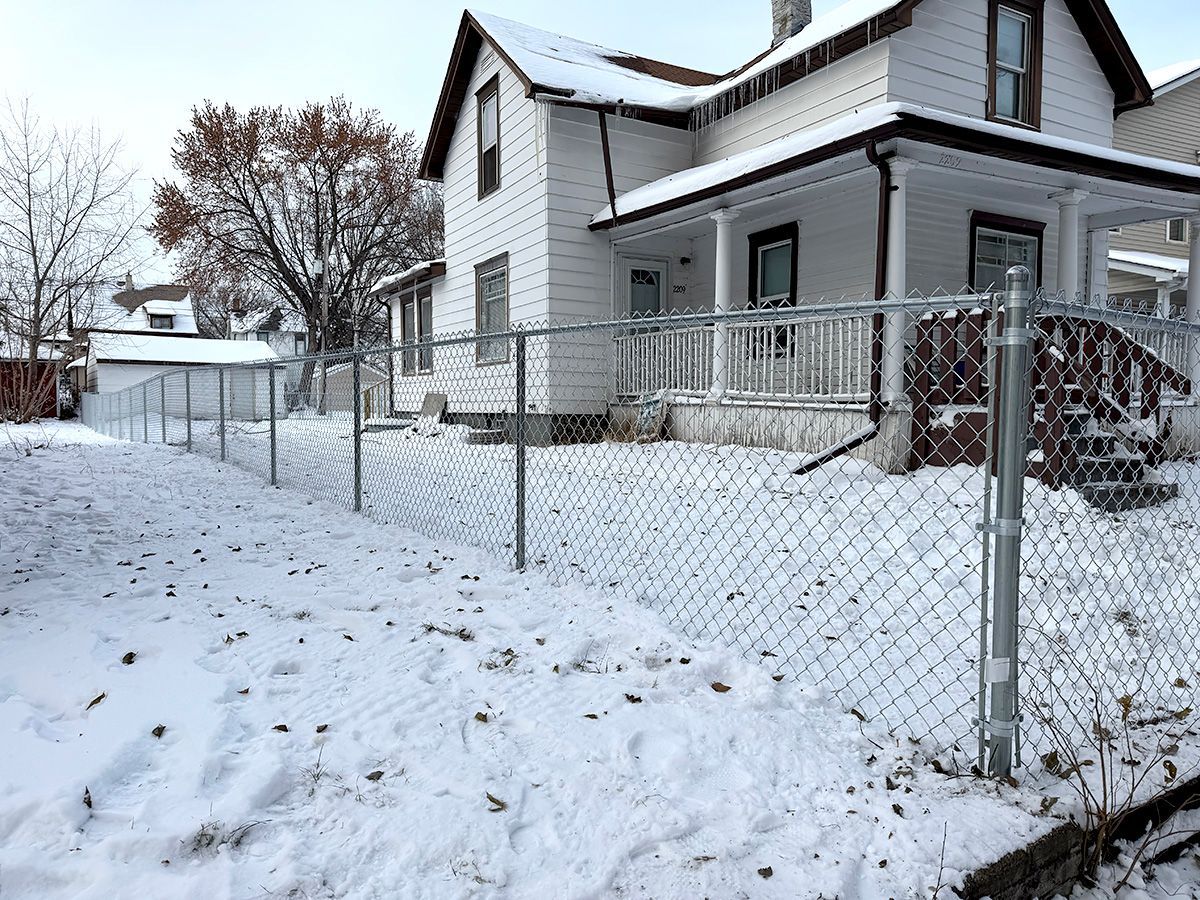 A white house with a front porch sits behind a chain-link fence on a snow-covered yard.