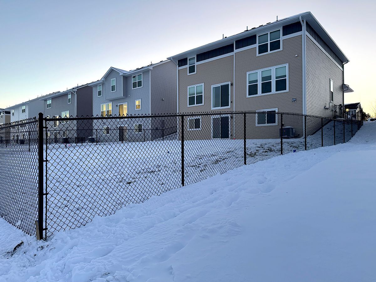 Tan and grey townhomes stand in a line behind a chain-link fence, with snow covering the ground and a clear sky above.