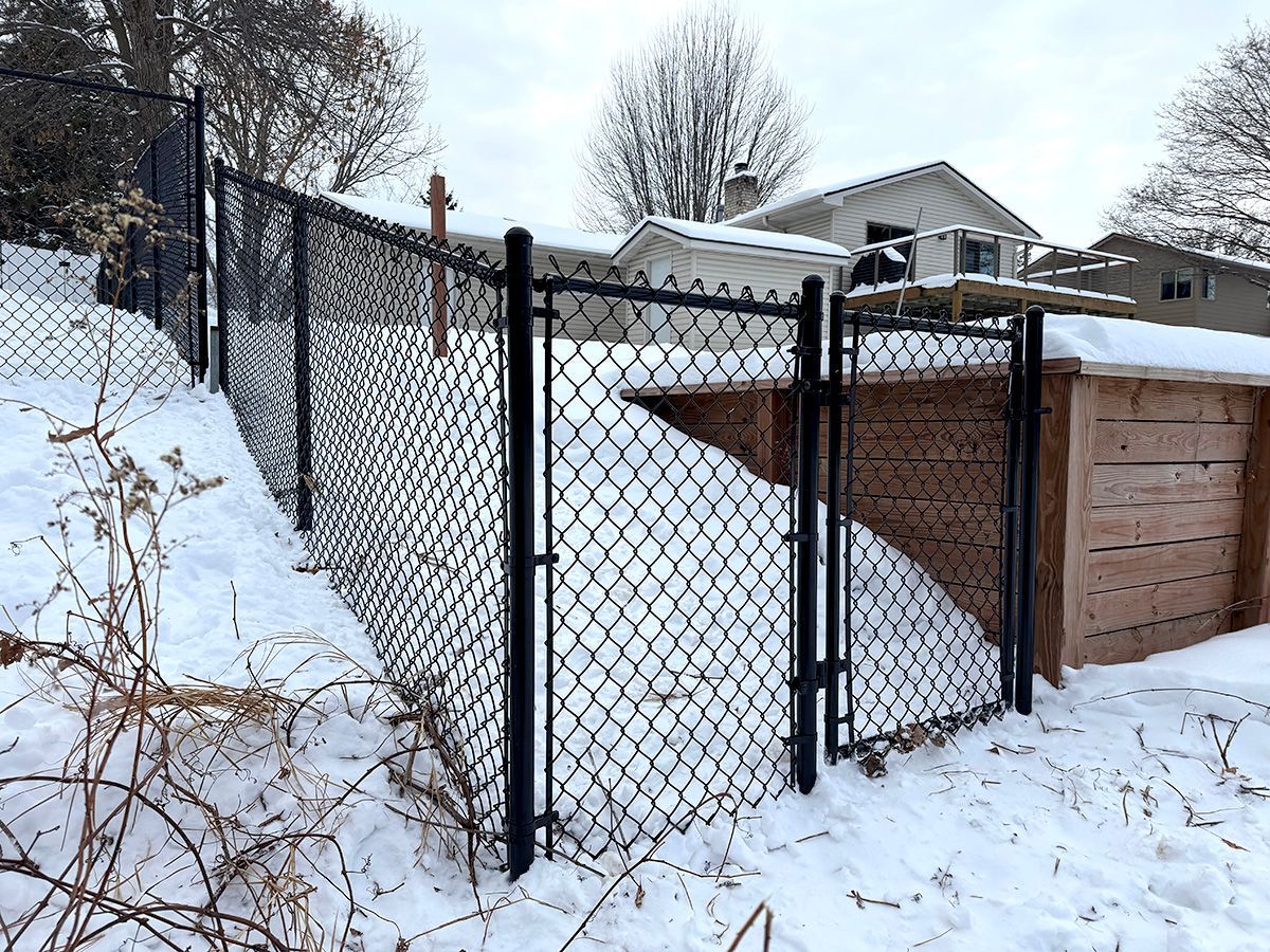 A black chain-link fence and a small gate enclose a snow-covered yard next to a wooden retaining wall.
