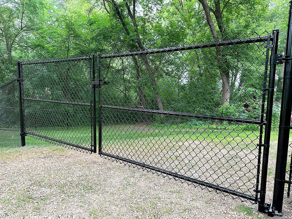 A black metal chain-link double gate stands closed, set against a background of lush green trees and a grassy lawn.
