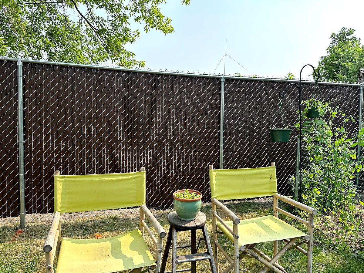Two yellow folding chairs sit on grass in front of a brown chain-link fence, with a small potted plant on a stool between.