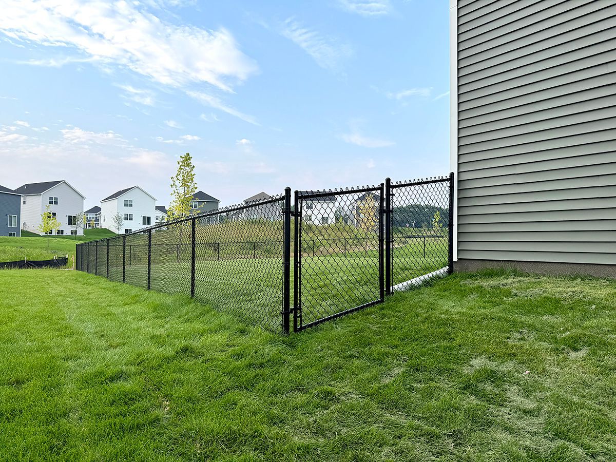 A black chain-link fence encloses a green grassy yard next to the side of a light gray house.