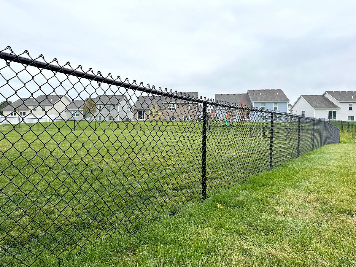 A black chain-link fence lines a green grassy field with suburban houses in the background under a cloudy sky.