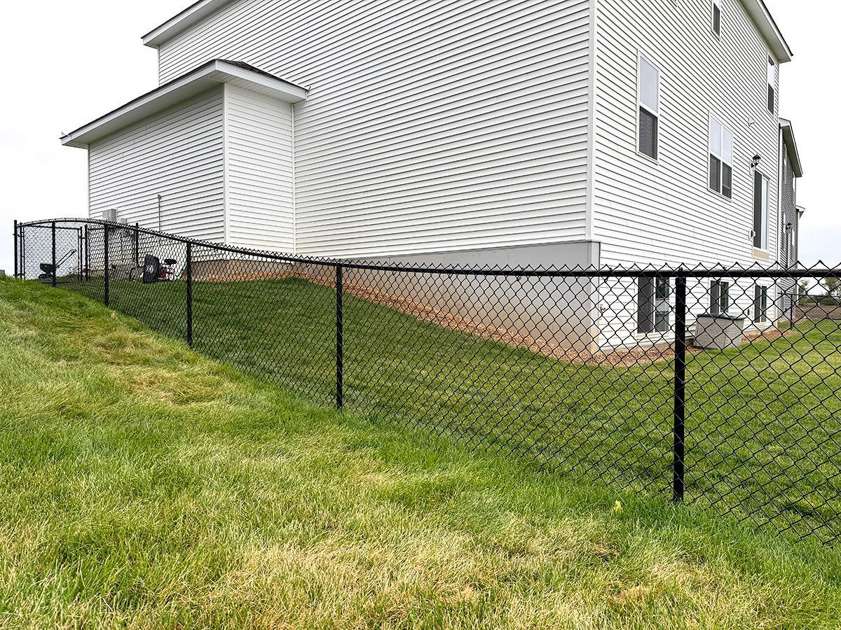 A black chain-link fence runs along a grassy slope beside the side of a white vinyl-sided house.