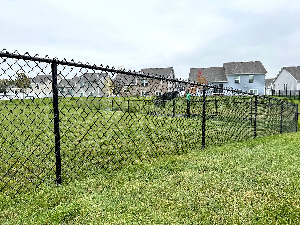 A black chain-link fence encloses a green grassy lawn in front of suburban houses under a cloudy sky.