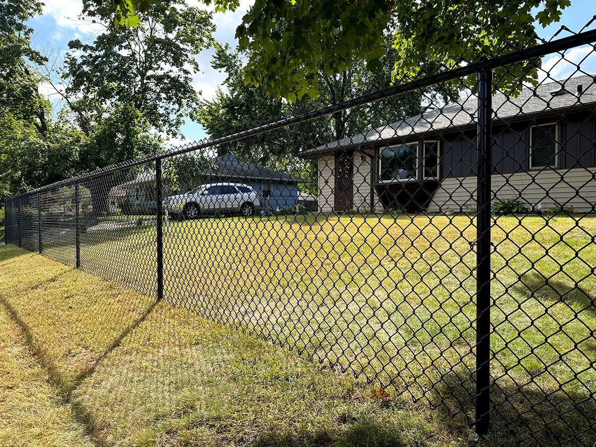 A black chain-link fence lines a lawn in front of a house, with a white SUV parked near a garage in the background.