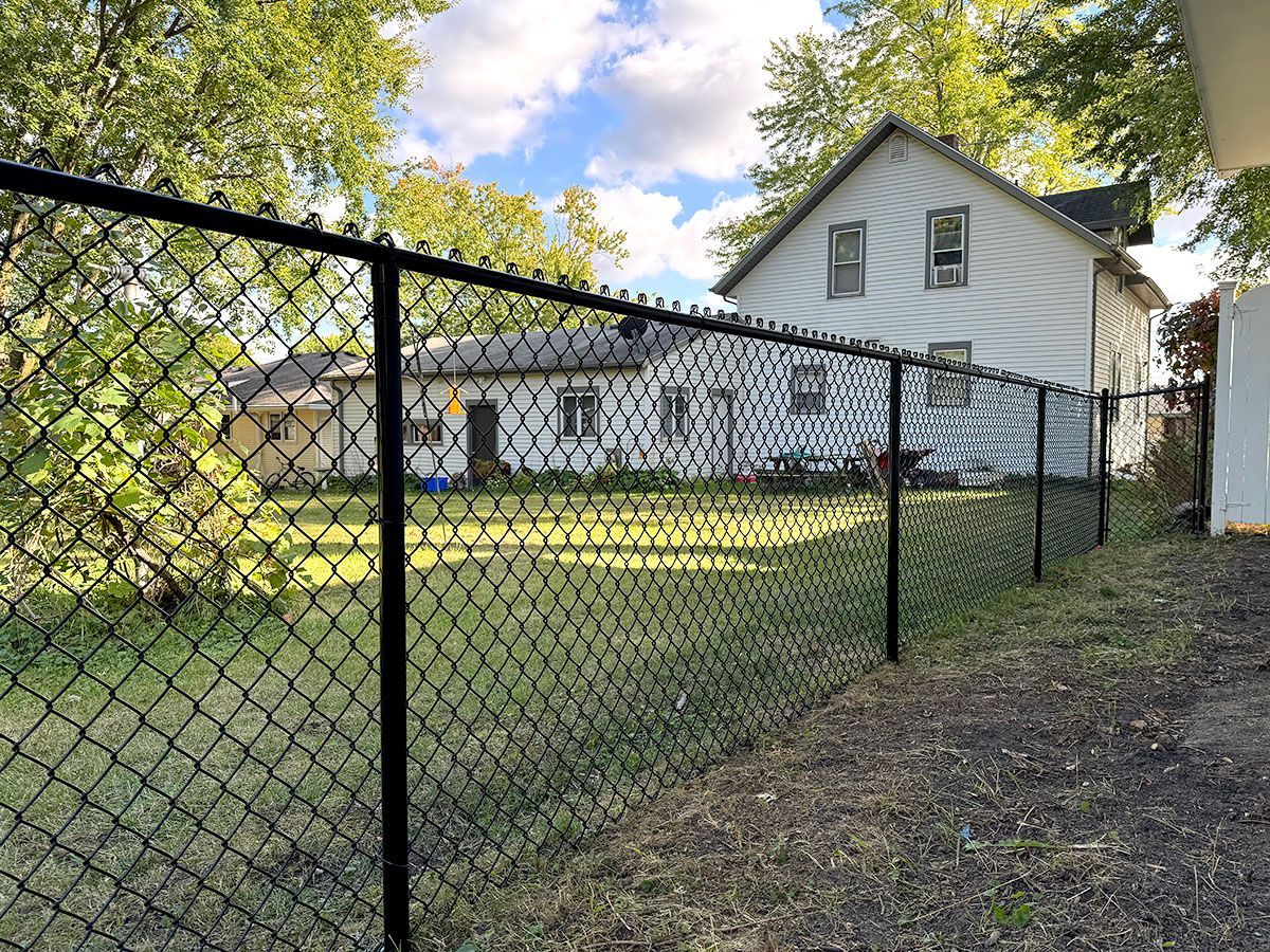 A black chain-link fence in the foreground, with a white house and green lawn in the backyard.
